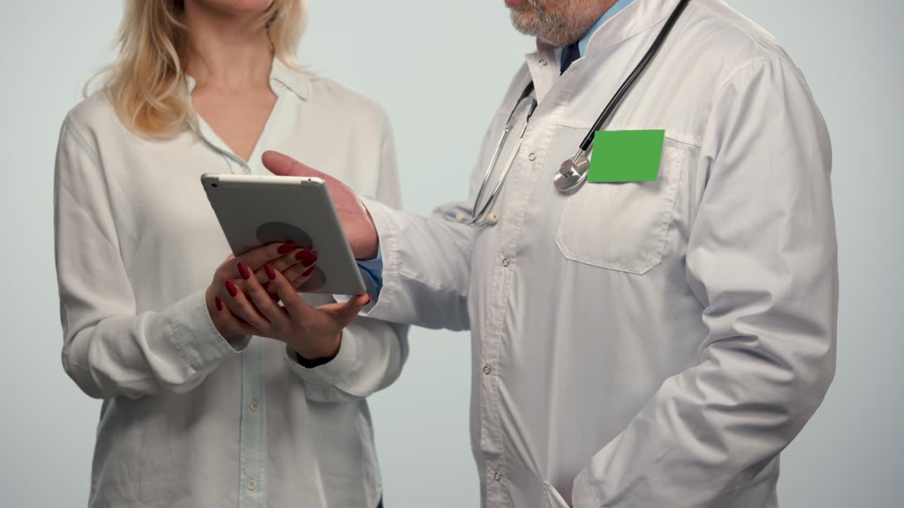 el médico de familia habla y muestra información en una tableta durante la consulta de una mujer paciente en un hospital. hombre médico en túnica médica blanca con insignia pantalla verde llave de croma. primer plano. cámara lenta