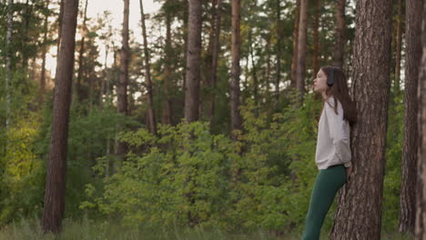 woman listens to music on headphones in forest. female leans on tree and ponders meaning of life during walk at sunset. calm atmosphere and feeling of solitude