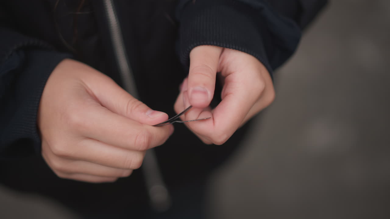 Primo piano di mani che fissano un fermaglio per capelli al crepuscolo, donna asiatica che sistema una piccola chiusura nera contro una giacca umida mentre le luci della città brillano sullo sfondo, movimenti concentrati mostrano determinazione e silenziosa resilienza.