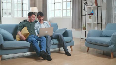 portrait of asian gentle gay couple using laptop computer, while sitting on a couch in cozy stylish apartment. adult boyfriends online shopping on internet, watching funny videos on streaming service