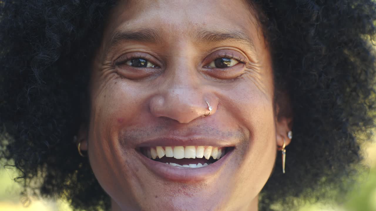 Homem africano sorrindo em uma gargalhada em um parque ensolarado, mostrando um piercing no nariz e vários brincos.
