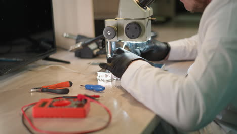 a close-up view of a technician in a white lab coat and black hand gloves meticulously testing a circuit under a microscope using a voltmeter in his laboratory, with various tools around