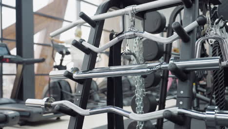 stacked layers of sleek chrome cable machine handles and attachments on sturdy rack in bright gym with blurred windows and cardio equipment in background showcasing modern fitness setup