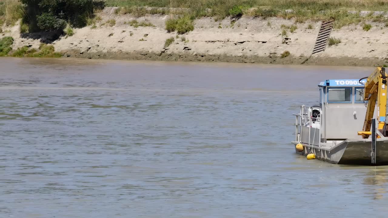 A small boat equipped with an excavator navigates a calm river near a sandy riverbank.