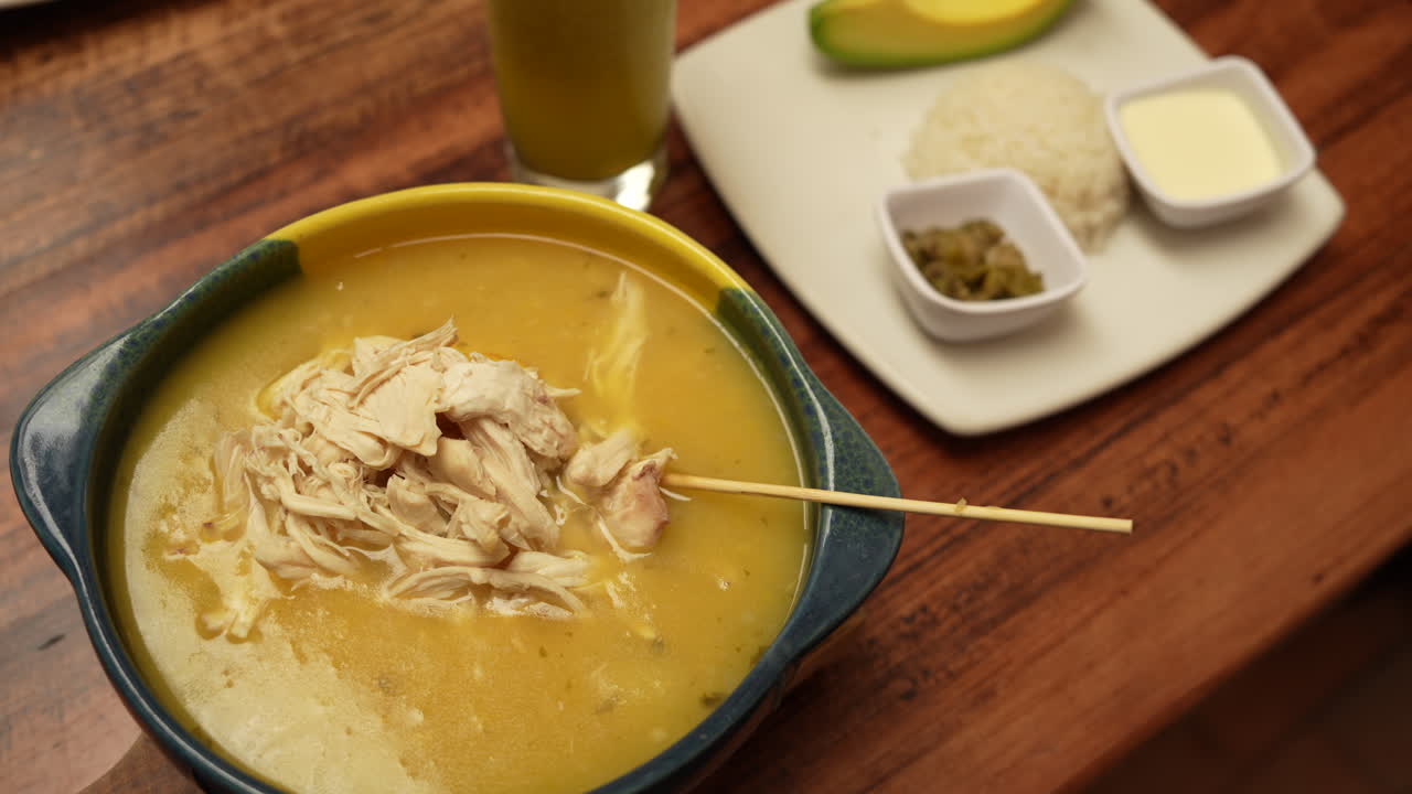 Delicious ajiaco soup in earthy bowl with shredded chicken, served with rice, avocado, and sauces on wooden table. Warm lighting enhances authentic Colombian dining atmosphere