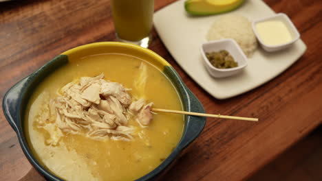delicious ajiaco soup in earthy bowl with shredded chicken, served with rice, avocado, and sauces on wooden table. warm lighting enhances authentic colombian dining atmosphere