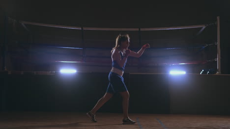 A-beautiful-woman-conducts-a-shadow-fight-practicing-technique-and-speed-of-strikes-while-training-hard-for-future-victories.-Dark-gym-background.-steadicam-shot