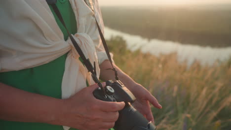 close up partial view of woman in green dress gently removing lens cover from camera while preparing to take photo, soft golden hour light casting warm glow across hands and natural field background