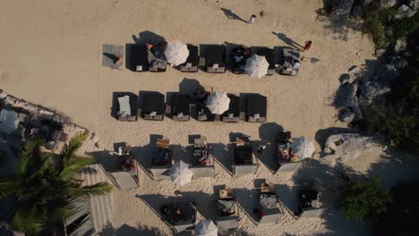 Toma-Aérea-De-Arriba-Hacia-Abajo-De-Personas-Relajándose-En-Una-Playa-Privada-De-Arena-Con-Bahía-A-La-Luz-Del-Sol---México,-Tulum---Vista-Ascendente-Hacia-Abajo