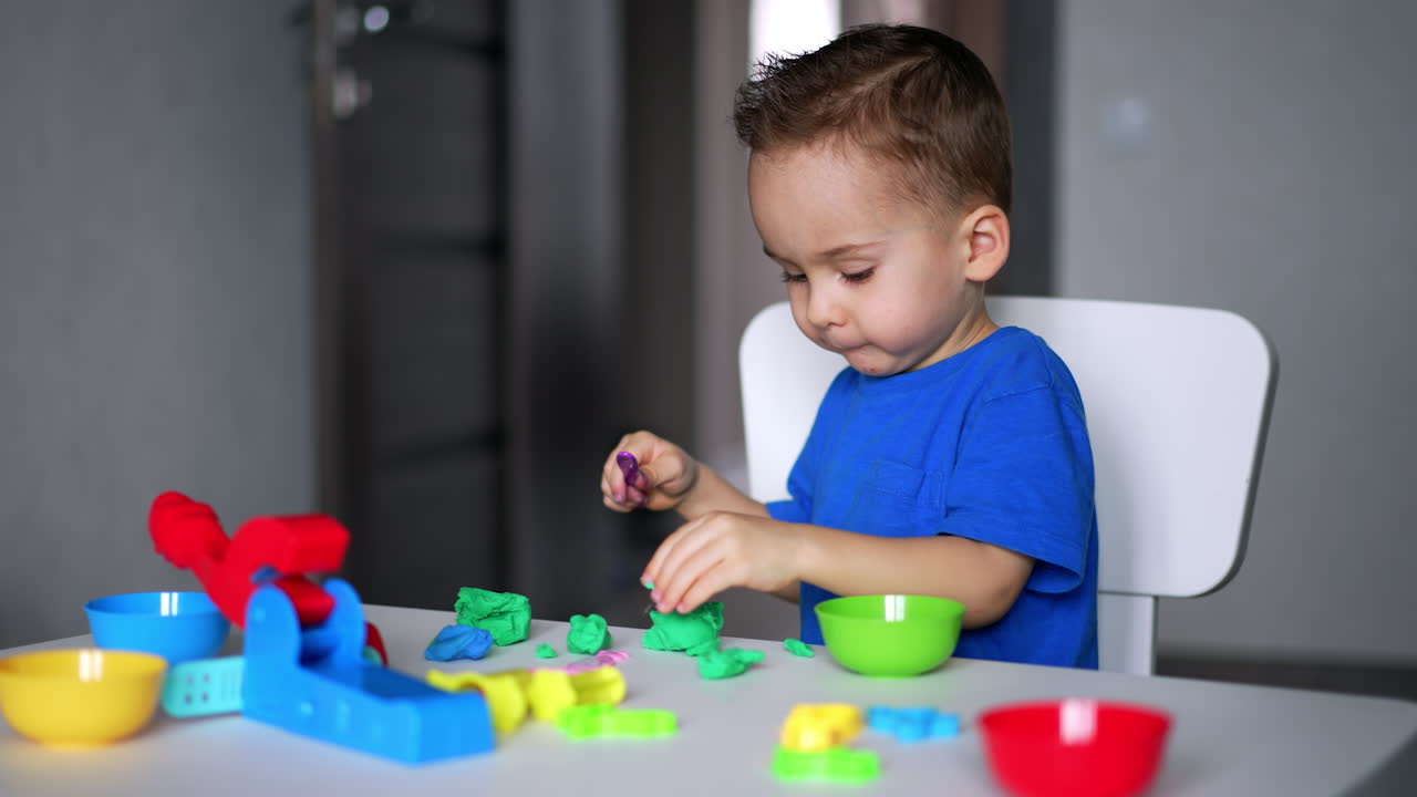 Cute Caucasian baby boy sits at desk playing with modeling dough. Focused kid cuts the plasticine with a stick.