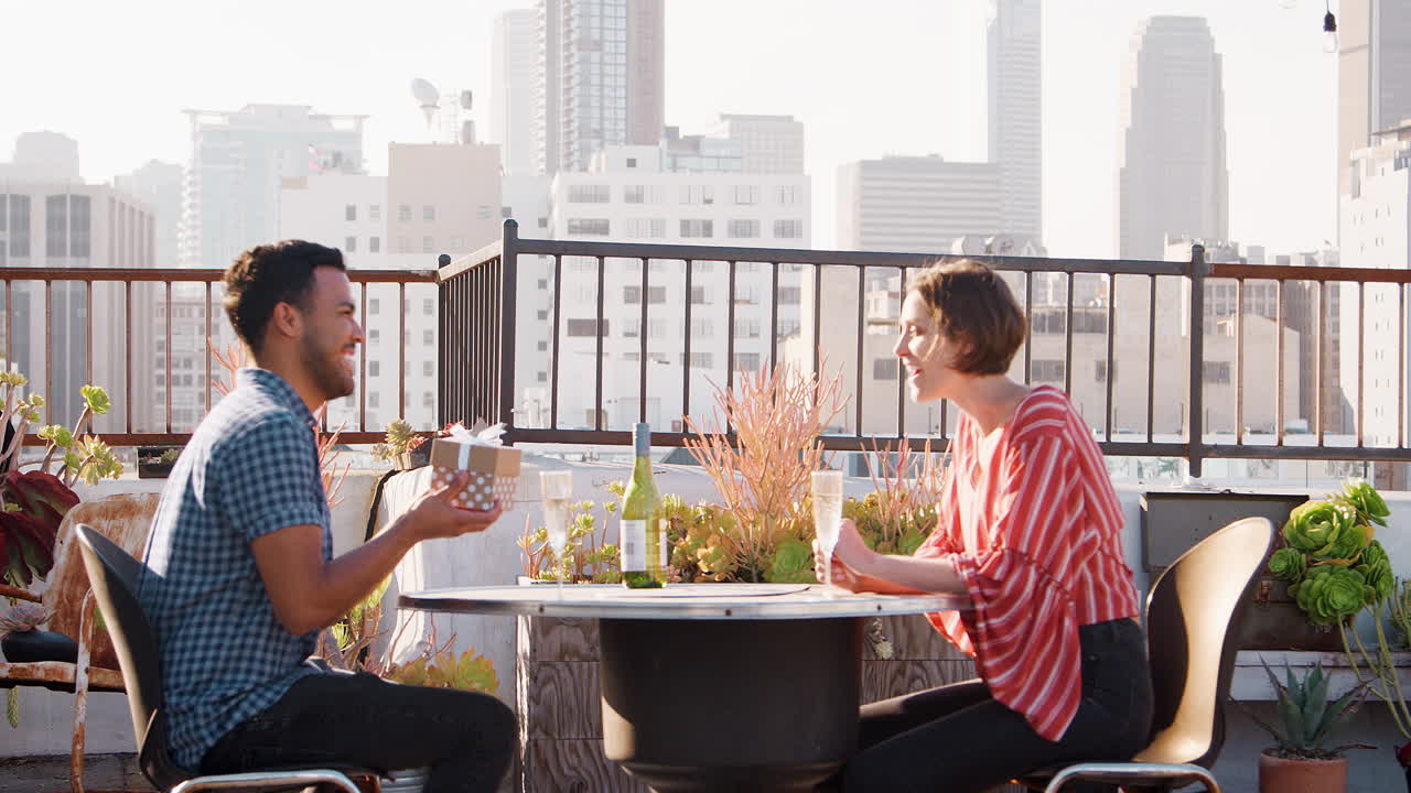 hombre dando regalo a la mujer mientras celebran en la terraza de la azotea con el horizonte de la ciudad en el fondo