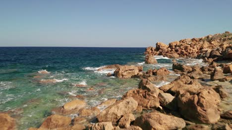 Toma-Aérea-De-Una-Costa-Con-Hermosas-Aguas-Turquesas-Del-Mar-Mediterráneo-Salpicando-En-Las-Rocas-De-Color-Rojo