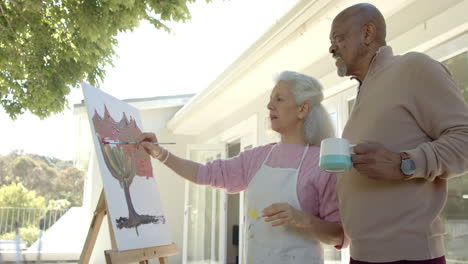 Happy-senior-biracial-couple-painting-and-drinking-coffee-at-balcony-at-home,-slow-motion