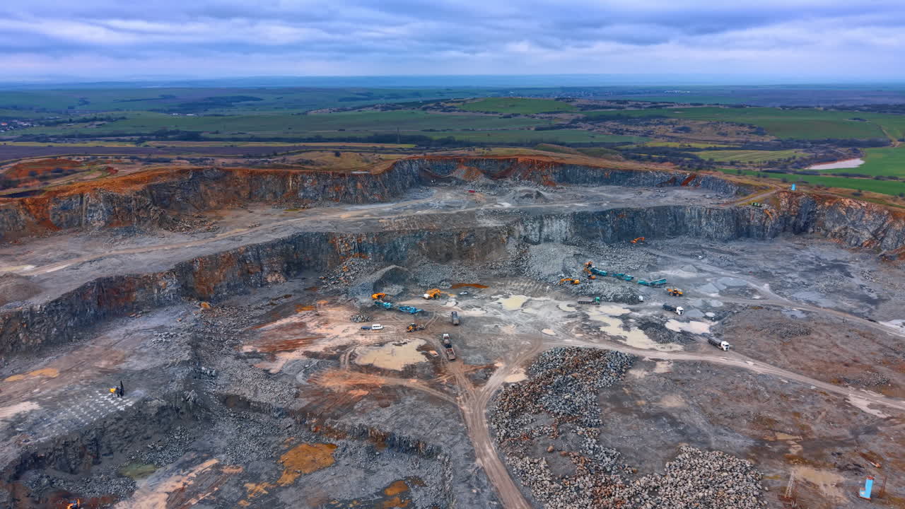 Panoramic aerial view of large scale industrial stone quarry. Wide panoramic shot of an active quarry landscape under a cloudy sky with green fields in the background