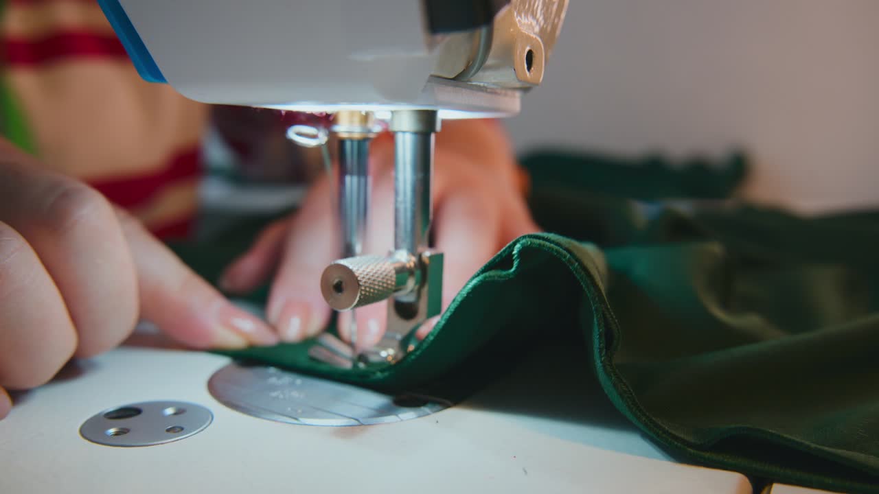 Hands guide green fabric under sewing machine needle. Close-up shows steady stitching motion. Fabric moves smoothly with precise control. Ideal for fashion, crafting, or textile industry projects