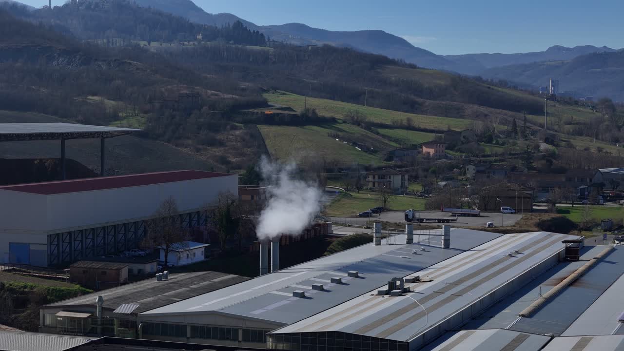 Bacalarından duman bulutları yükselen bir fabrika kompleksinin, parlak bir gökyüzü altında manzaralı dağlık bir peyzaj fonunda çekilmiş hava görüntüsü
