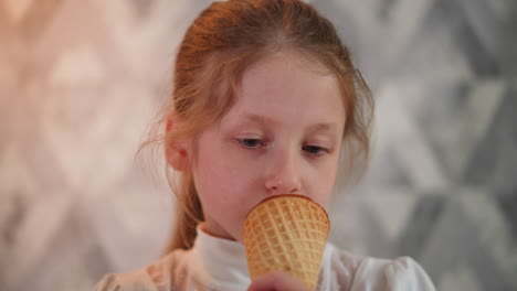 pretty young girl wearing white top casually eating ice cream cone against patterned wall background with soft lighting, focused expression enjoying sweet treat in relaxed indoor environment