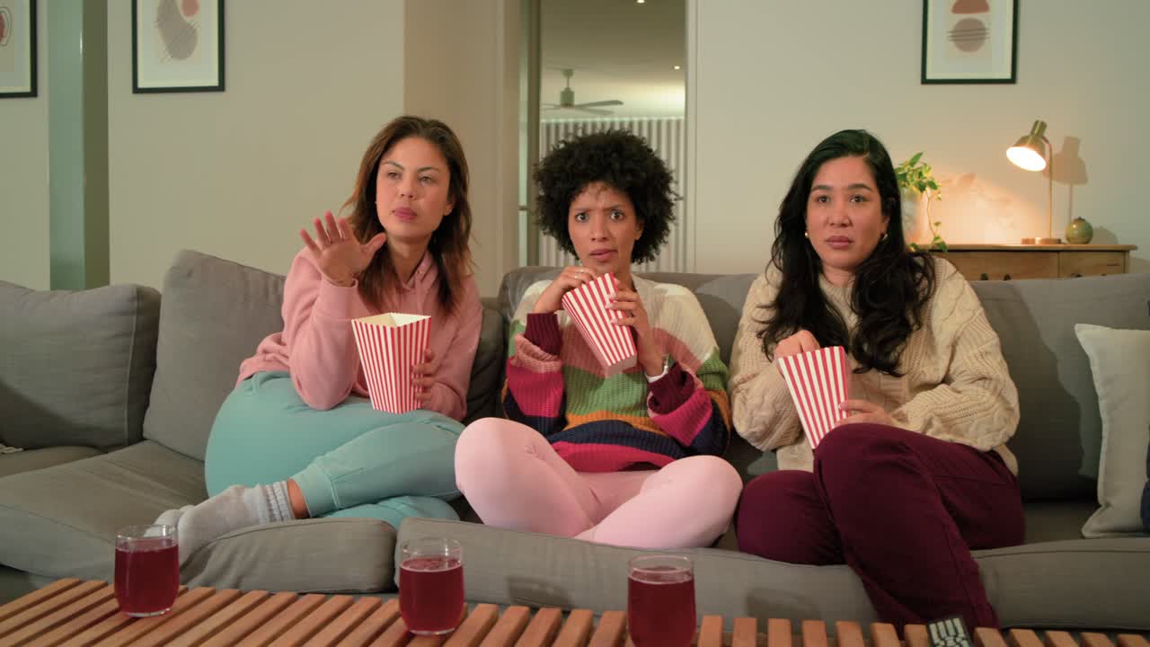 Tre amiche che mangiano popcorn da contenitori a righe su un divano, che reagiscono sorprese a ciò che vedono sullo schermo