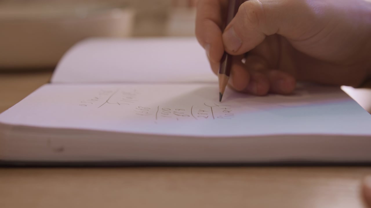 Close up of hand writing mathematical equations in pencil on white notebook paper showing detailed arithmetic calculations. scene features soft lighting and natural indoor setting