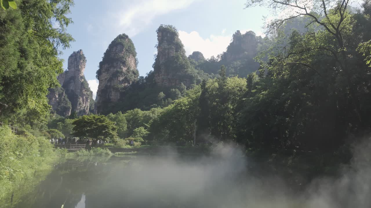Rừng sương mù với sương mù thấp ở Trương Gia Giới cùng những đám mây thấp di chuyển qua những tán cây xanh cận nhiệt đới rậm rạp, góc máy nghiêng lên từ ao đến các đỉnh tháp