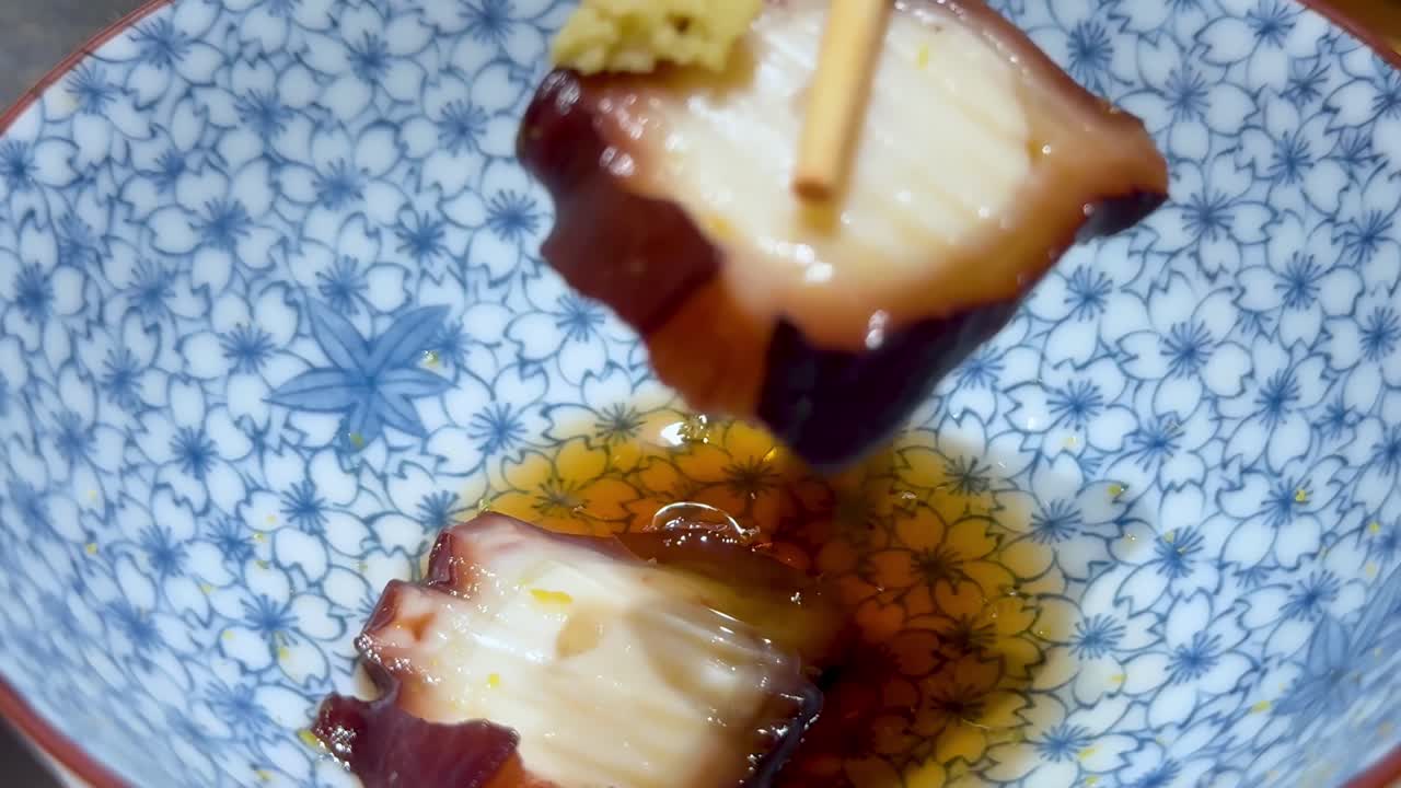 Close-up of chopsticks picking up octopus with wasabi in a blue patterned bowl.