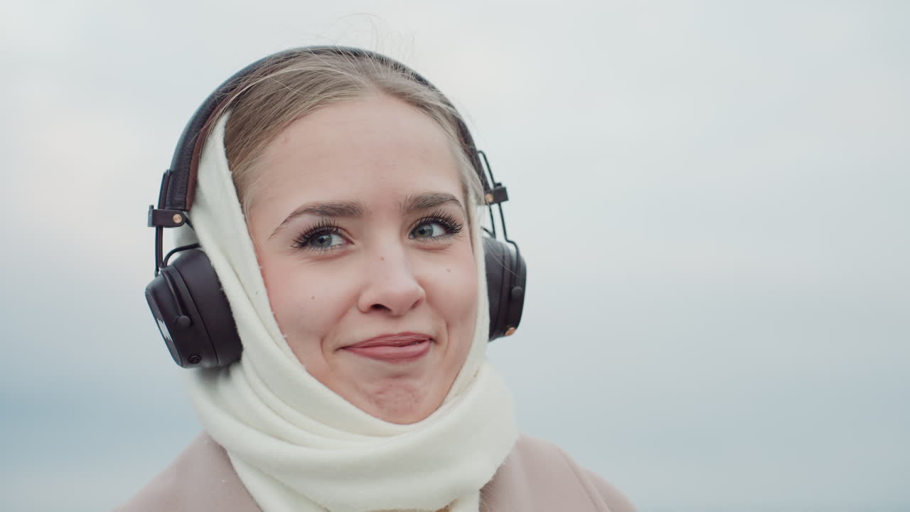 Ritratto invernale introspettivo. Donna serena con cuffie all'aperto. Donna con cuffie che guarda pensierosa il lungomare. Primo piano di donna in abbigliamento invernale con colori tenui e un'atmosfera riflessiva.