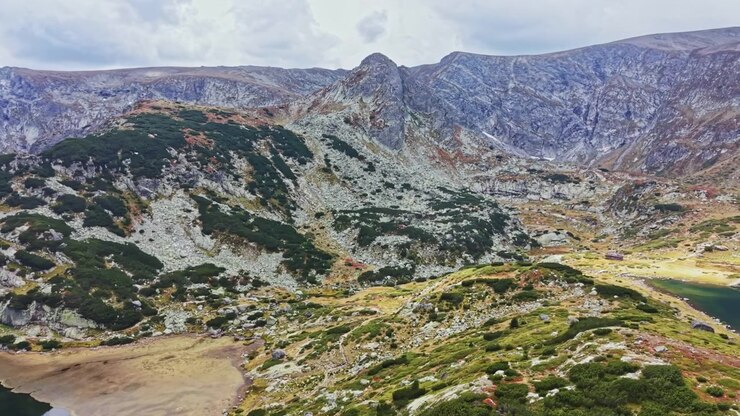 Paesaggio montano con terreno roccioso, arbusti verdi e un piccolo lago sotto un cielo nuvoloso in un contesto naturale