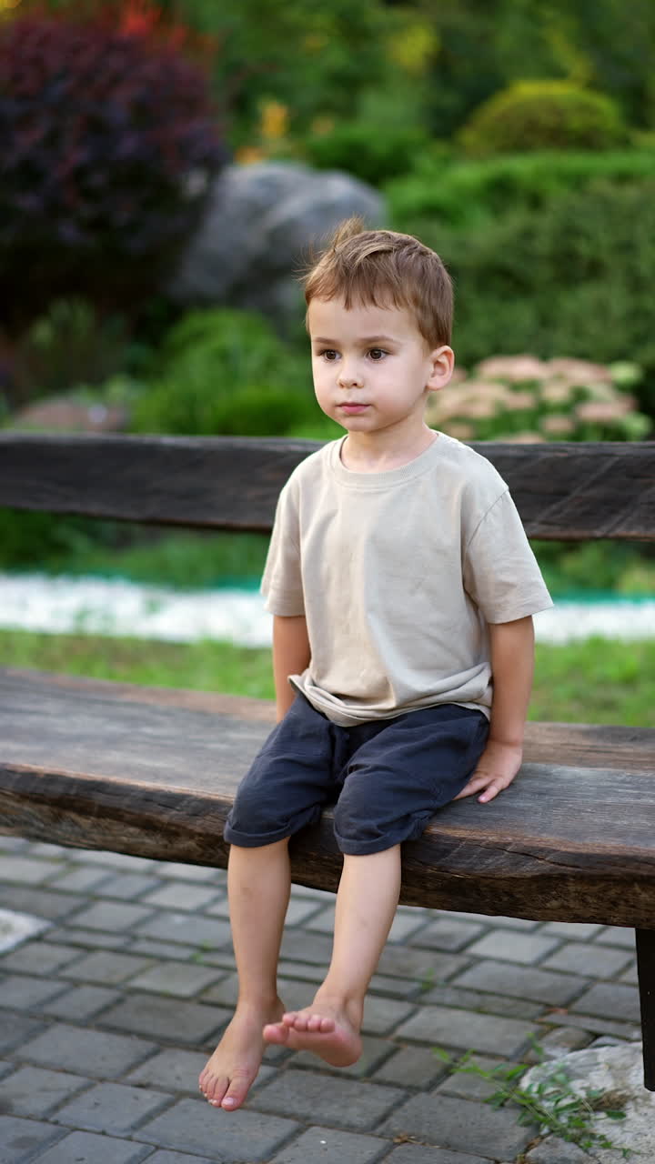 Cute baby sitting barefoot on the wooden bench with his feet up. Charming Caucasian toddler outdoors in summer. Vertical video.