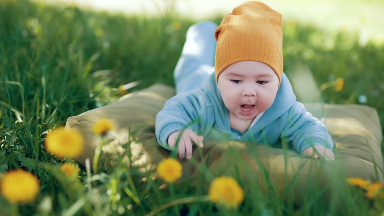 Active baby boy lies outdoors in green grass. Kid claps hands on the pillow and makes faces funnily.