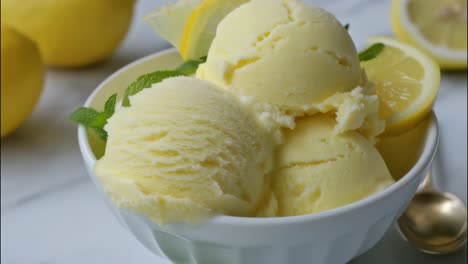 Scoops of refreshing lemon ice cream, garnished with mint and lemon slices, sit in a white bowl, ready to be enjoyed, with whole lemons in the soft focus background