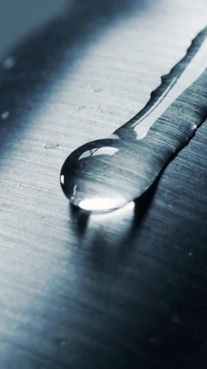 Vertical video: Sliding clear droplet tracing rivulet on sloped brushed metal, shifting highlights