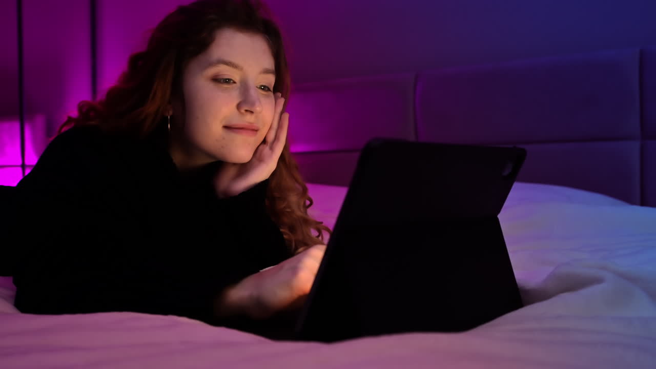 Young ginger hair girl browsing her tablet at night in the bedroom. Colorful light on background