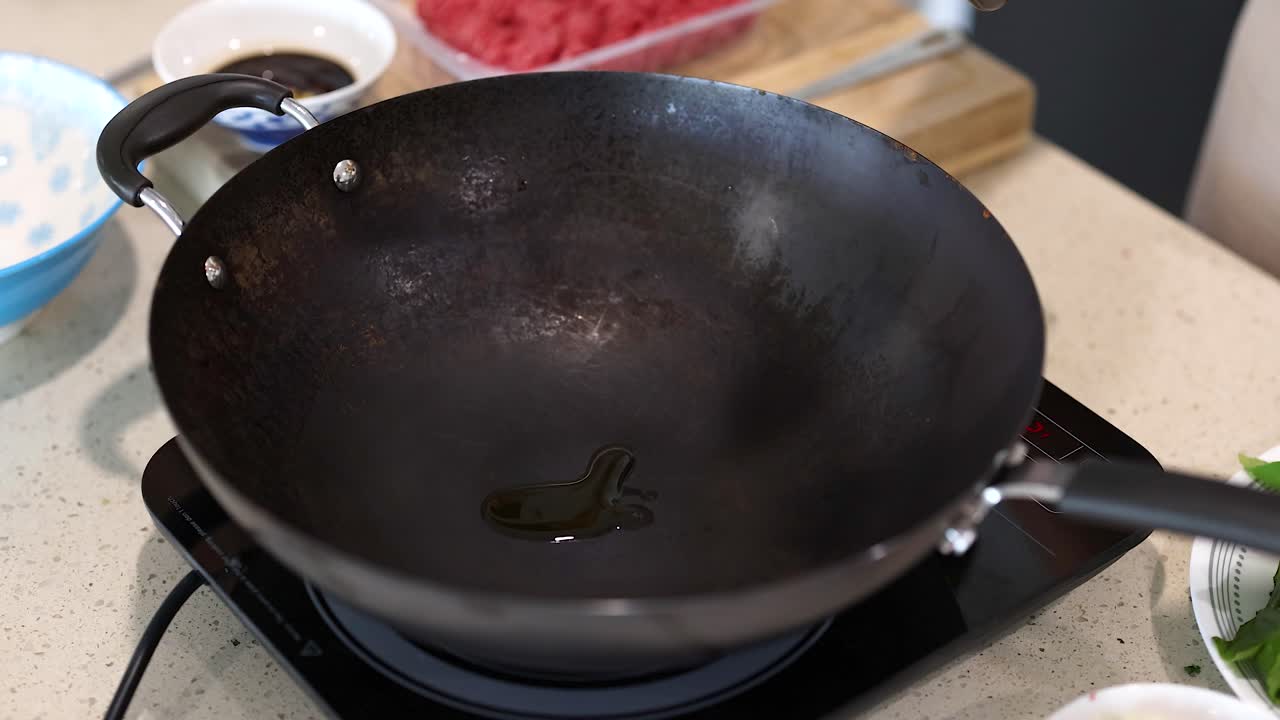 Oil is poured into a heated wok on a kitchen stove, preparing for cooking. Bright lighting highlights the kitchen setting