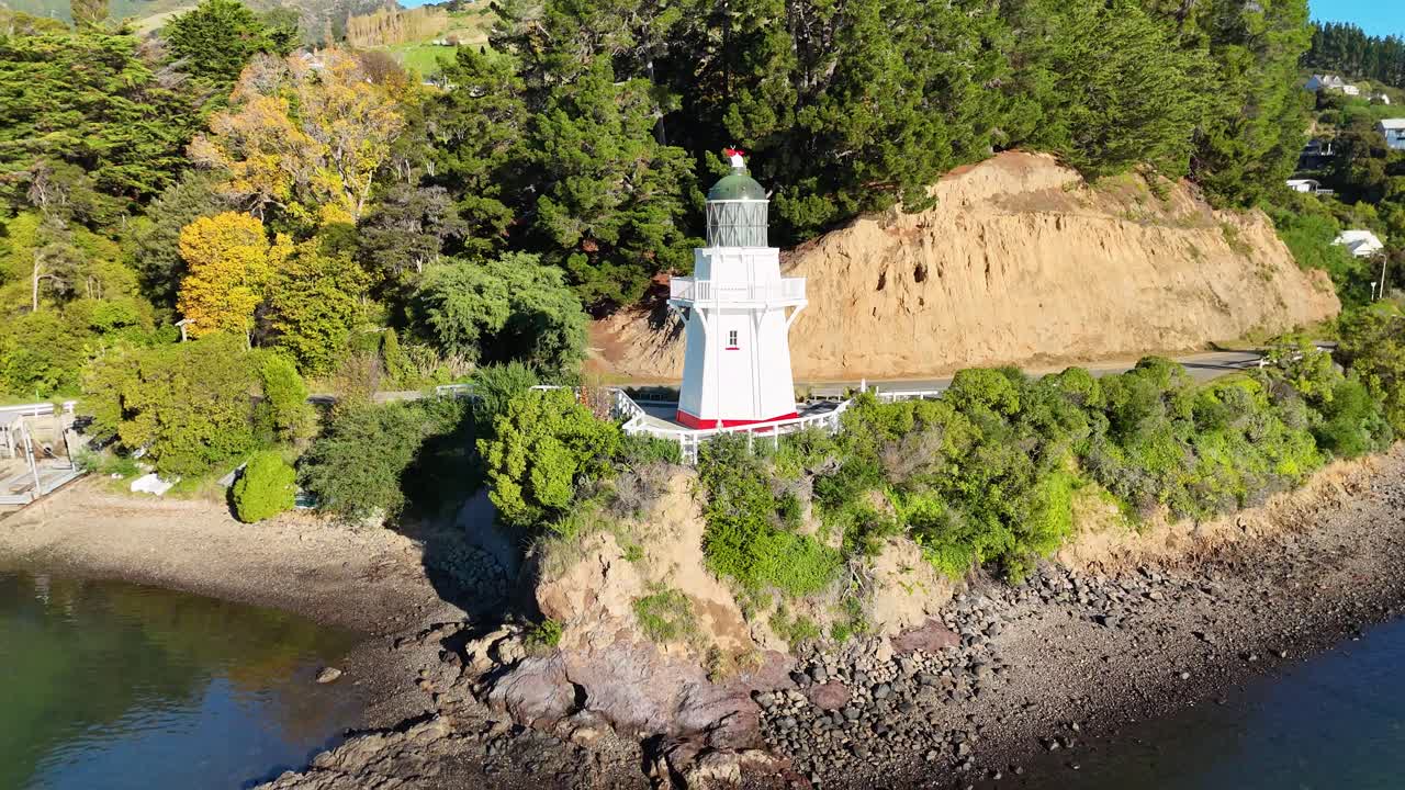 Aerial footage captures the scenic Akaroa Lighthouse surrounded by lush greenery and coastal waters in bright daylight