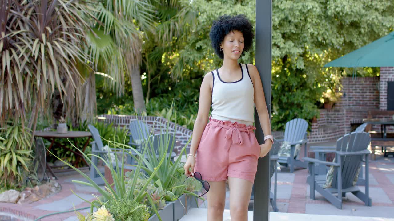 Mujer afroamericana adulta notando la cámara, caminando por el patio en pantalones cortos y gafas de sol, posando junto a un poste