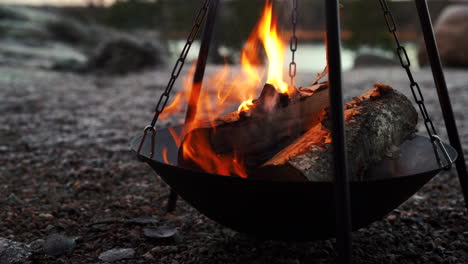 big wooden fire logs burning on stone gravel, camp fire in front of lake