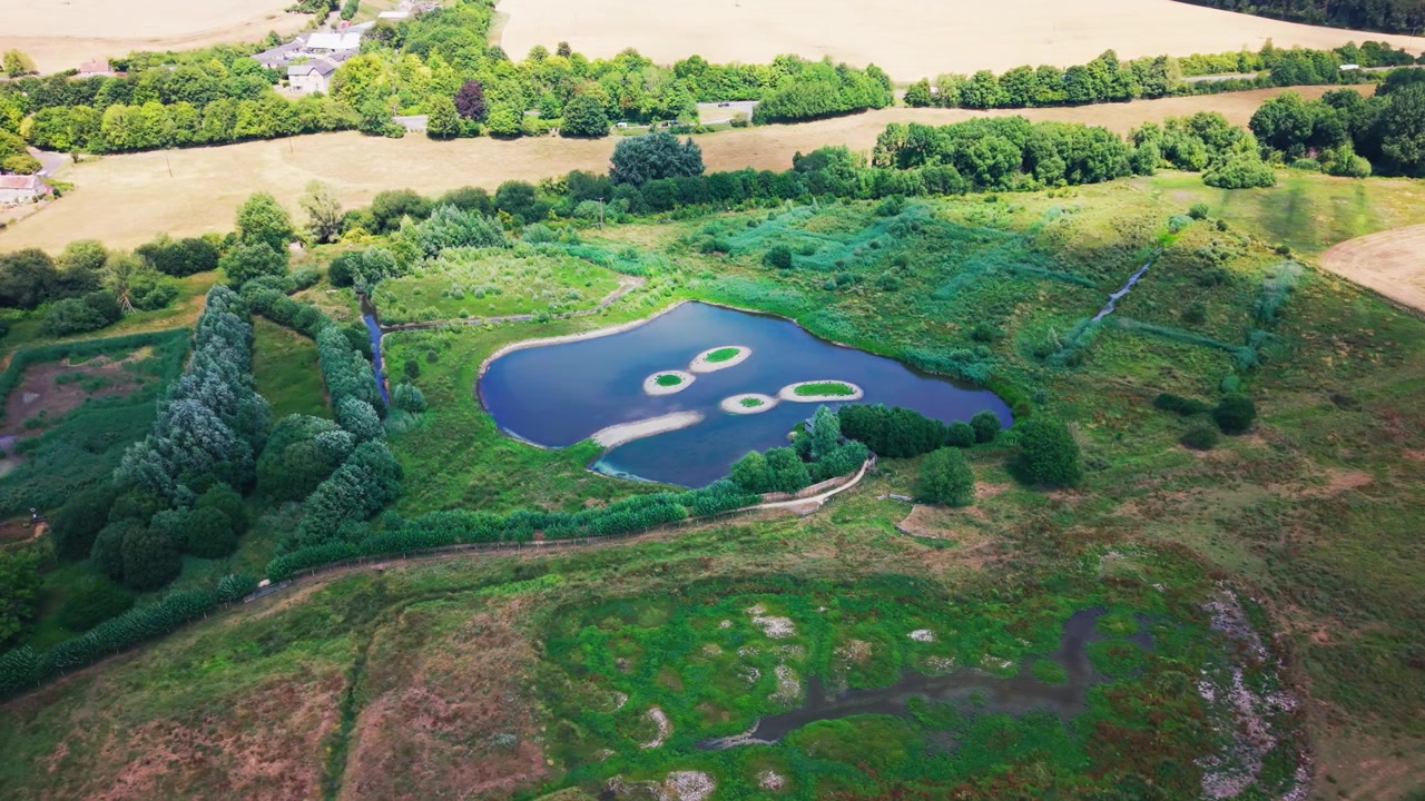 Weitwinkel-Drohnenaufnahme aus der Luft mit Blick auf eine renaturierte Süßwasser-Feuchtgebietslagune mit Kies-Nistinseln, Schilfgürteln, Grasland und umgebendem Waldgebiet im Naturschutzgebiet Langford Lakes in Wiltshire.