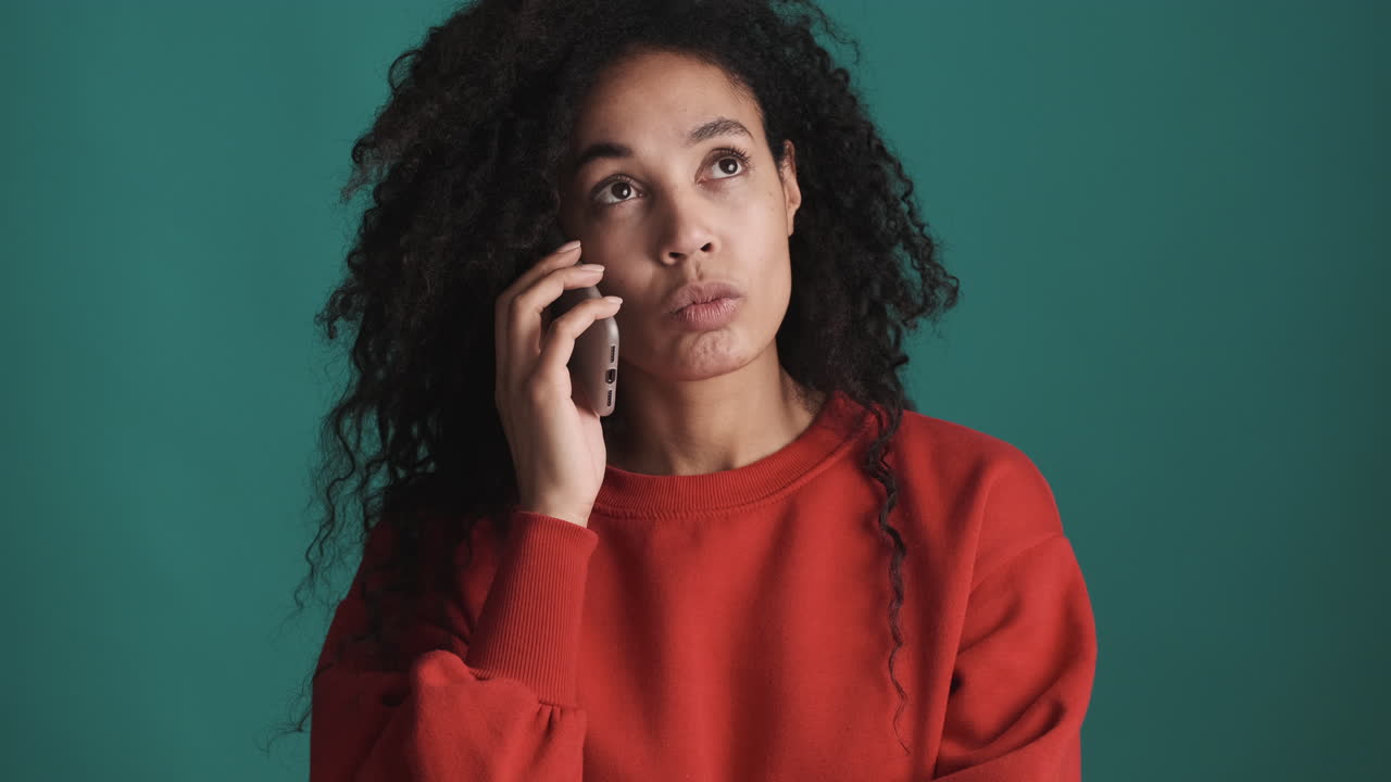 una mujer afroamericana pensativa usando un teléfono inteligente sobre un fondo azul.