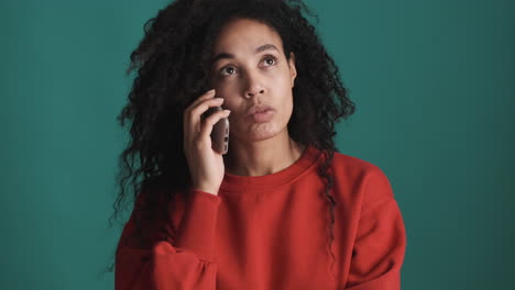 una mujer afroamericana pensativa usando un teléfono inteligente sobre un fondo azul.