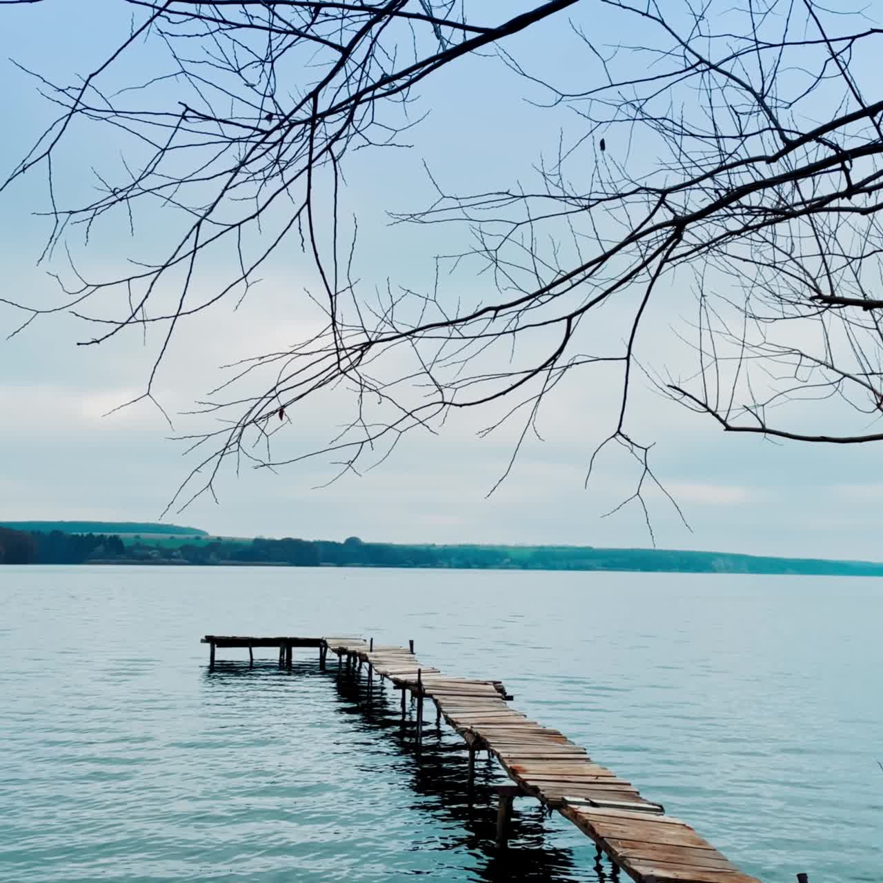 Meditative scenery of river with long wooden bridge on. Tree leafless branches hanging above. Hazy dark waterfront at backdrop