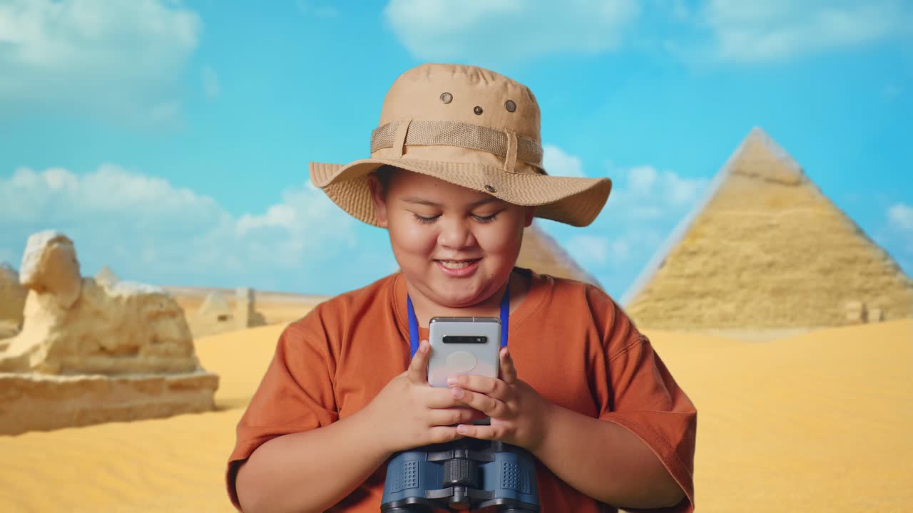 Asian Boy With A Hat And Binoculars Looking At Smartphone And Smiling While Traveling In Giza Pyramid. Boy Researcher Examines Something, Travel Adventure Concept, Close Up