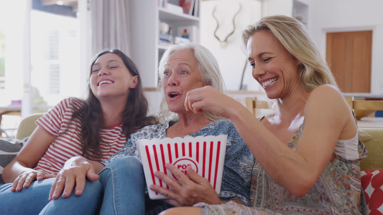 Mor med voksen datter og teenagedatterdatter spiser popcorn og ser film på sofaen derhjemme.