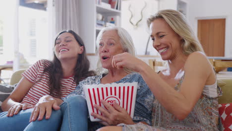 madre con hija adulta y nieta adolescente comiendo palomitas de maíz viendo una película en el sofá en casa
