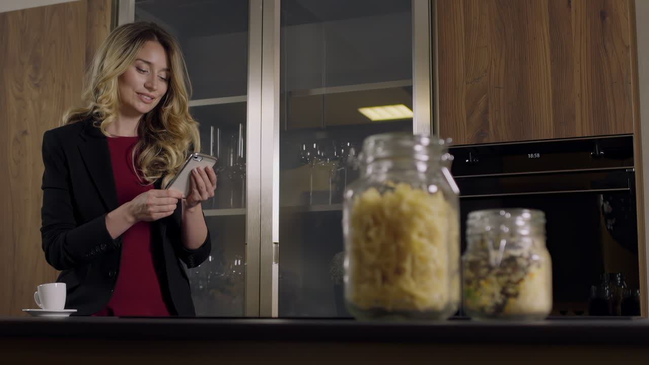 A woman dressed in professional attire is smiling as she texts on her phone in a stylish, modern kitchen. Jars of ingredients are on the counter, creating a cozy atmosphere