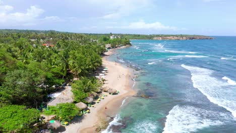 aerial shot over the najayo beach with a view of thatched houses on the shore of the beach on a clear afternoon, breeze hitting the palms, sea with waves