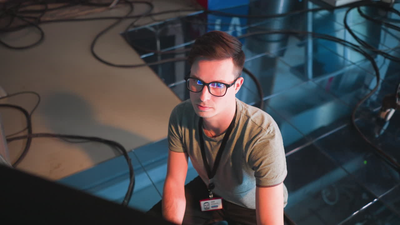 Person focused on computer monitor in TV studio. Wearing glasses and a lanyard, showing deep concentration while working in a modern tech environment. Background features cables and lighting