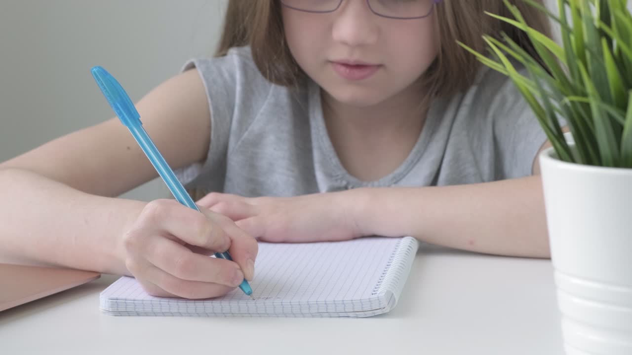 Close up kid's girl hand making notes in notebook at desk. Child girl holding pen doing homework