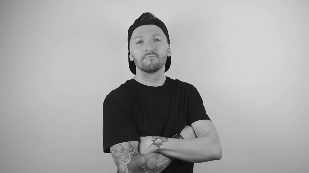 Black and white footage of a tattooed man with a sleeve crossing his arms and hands and nodding with approval and confidence in slow motion. Video is filmed on a white background that has shadows.