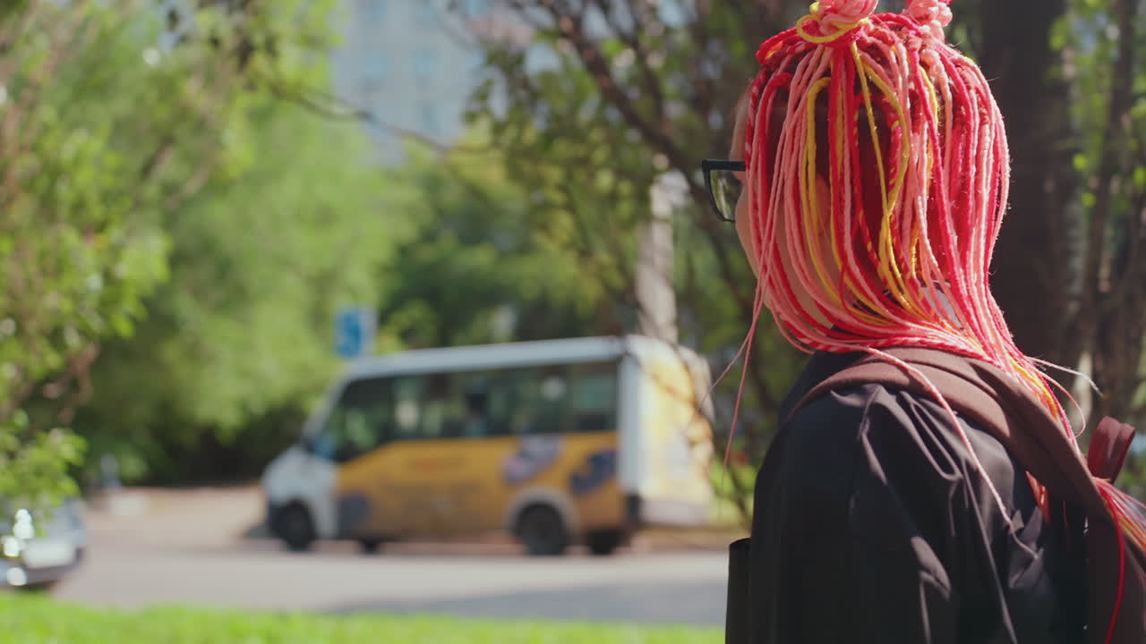 Una mujer con el pelo de colores observa los coches que pasan, una mujer junto al mar con el pelo llamativo se detiene a observar los vehículos que pasan, una mujer reflexiva con el pelo de color carmesí brillante está de pie junto a la carretera observando el paso de los vehículos