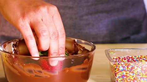 A close-up view of strawberries being dipped in chocolate and coated with colorful sprinkles.